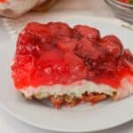 A slice of Pretzel Jello Salad dessert with a crumbly base, creamy middle layer, and a topping of glazed strawberries sits elegantly on a white plate.