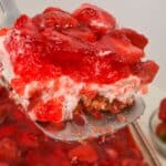 A close-up of a spatula lifting a slice featuring a crumbly pretzel crust, creamy layer, and glossy strawberry topping.