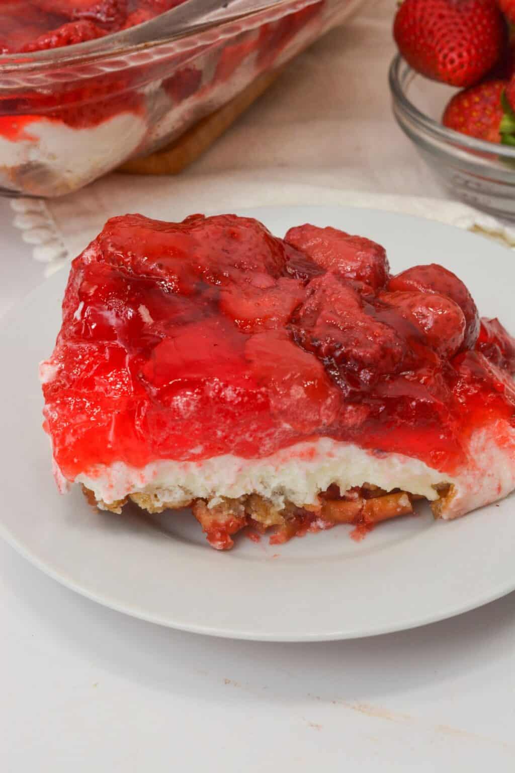 A slice of Pretzel Jello Salad with a crumbly base, creamy middle layer, and bright strawberry topping sits on a white plate.