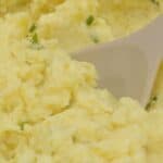 Creamy Boursin Mashed Potatoes with chopped green herbs mixed in, being stirred with a beige spoon.