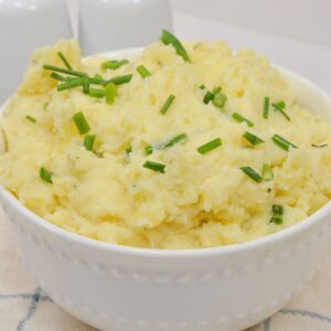 A white bowl filled with creamy Boursin Mashed Potatoes, garnished with chopped chives, placed on a light-colored surface.