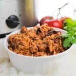 A white bowl filled with shredded Chipotle Slow Cooker Chicken sits in front of a Crock-Pot, with tomatoes and cilantro in the background.