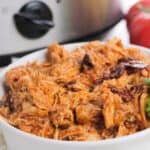 A white bowl filled with shredded Chipotle Slow Cooker Chicken sits in front of a slow cooker, with a tomato visible in the background.