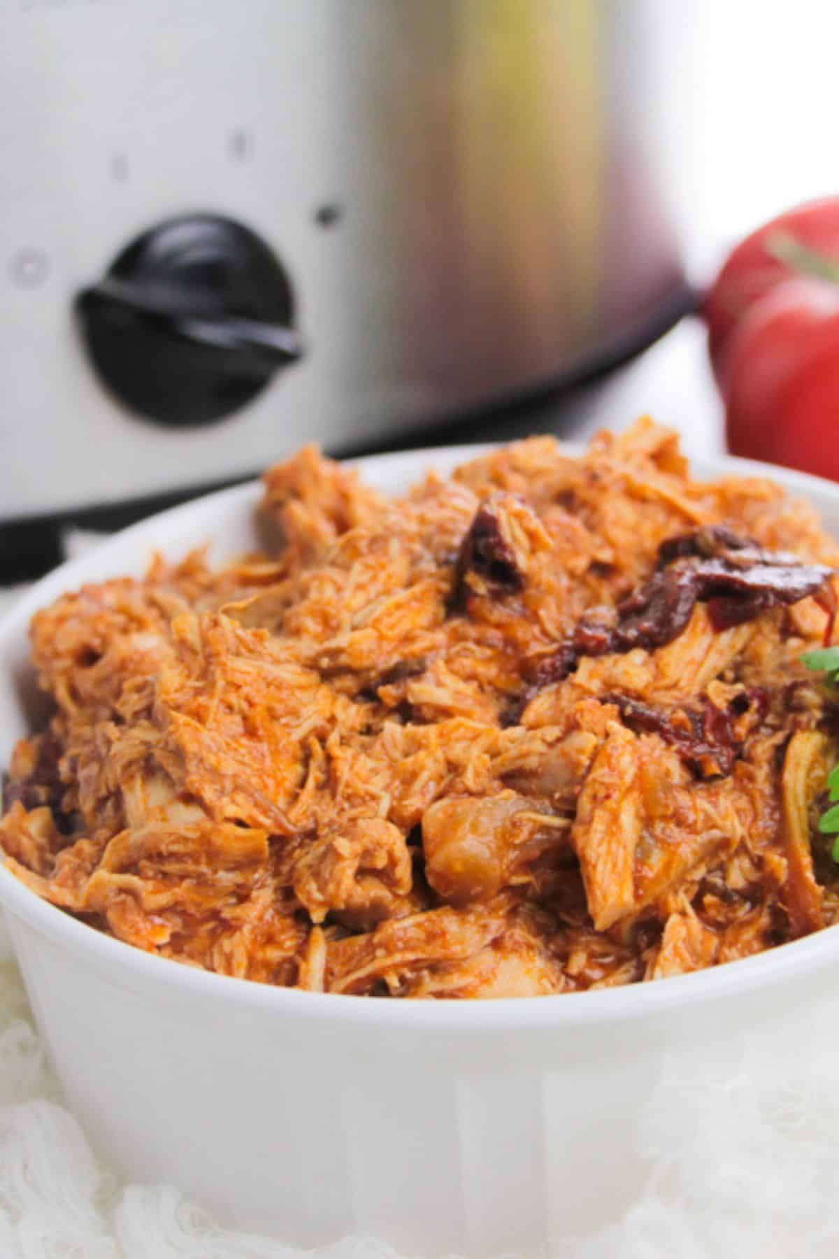 A white bowl filled with shredded Chipotle Slow Cooker Chicken sits in front of a slow cooker, with a tomato visible in the background.