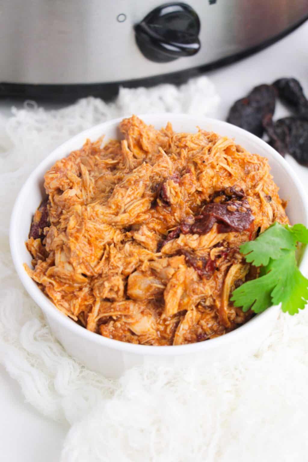 A white bowl filled with shredded Chipotle Slow Cooker Chicken, garnished with a sprig of cilantro, sits in front of a slow cooker and some dried peppers.