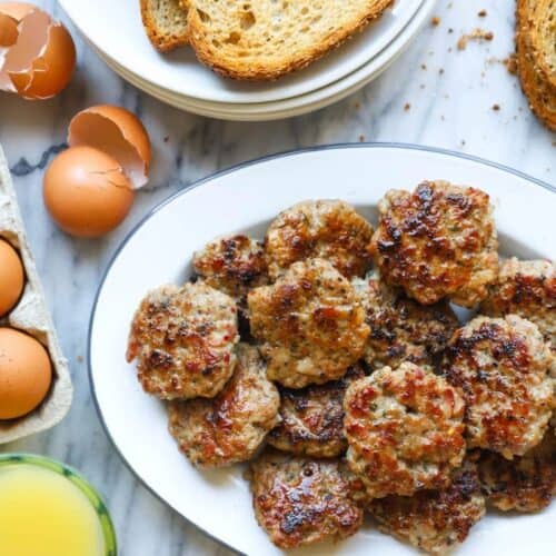 A white platter of cooked breakfast sausage patties on a marble surface, surrounded by slices of toast, whole eggs, cracked eggshells, and a glass of orange juice—a classic scene from delicious breakfast sausage recipes.