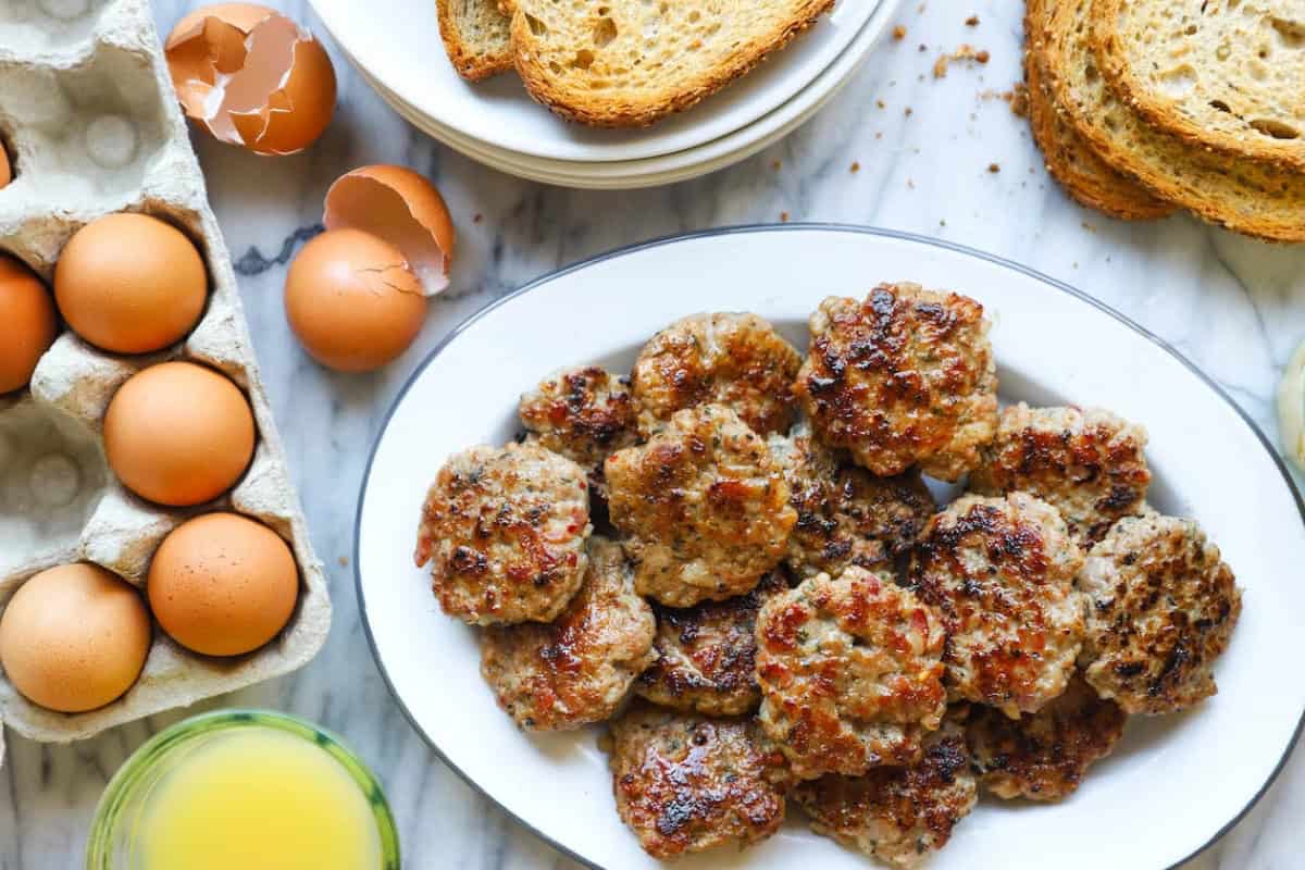 A white platter of cooked breakfast sausage patties on a marble surface, surrounded by slices of toast, whole eggs, cracked eggshells, and a glass of orange juice.
