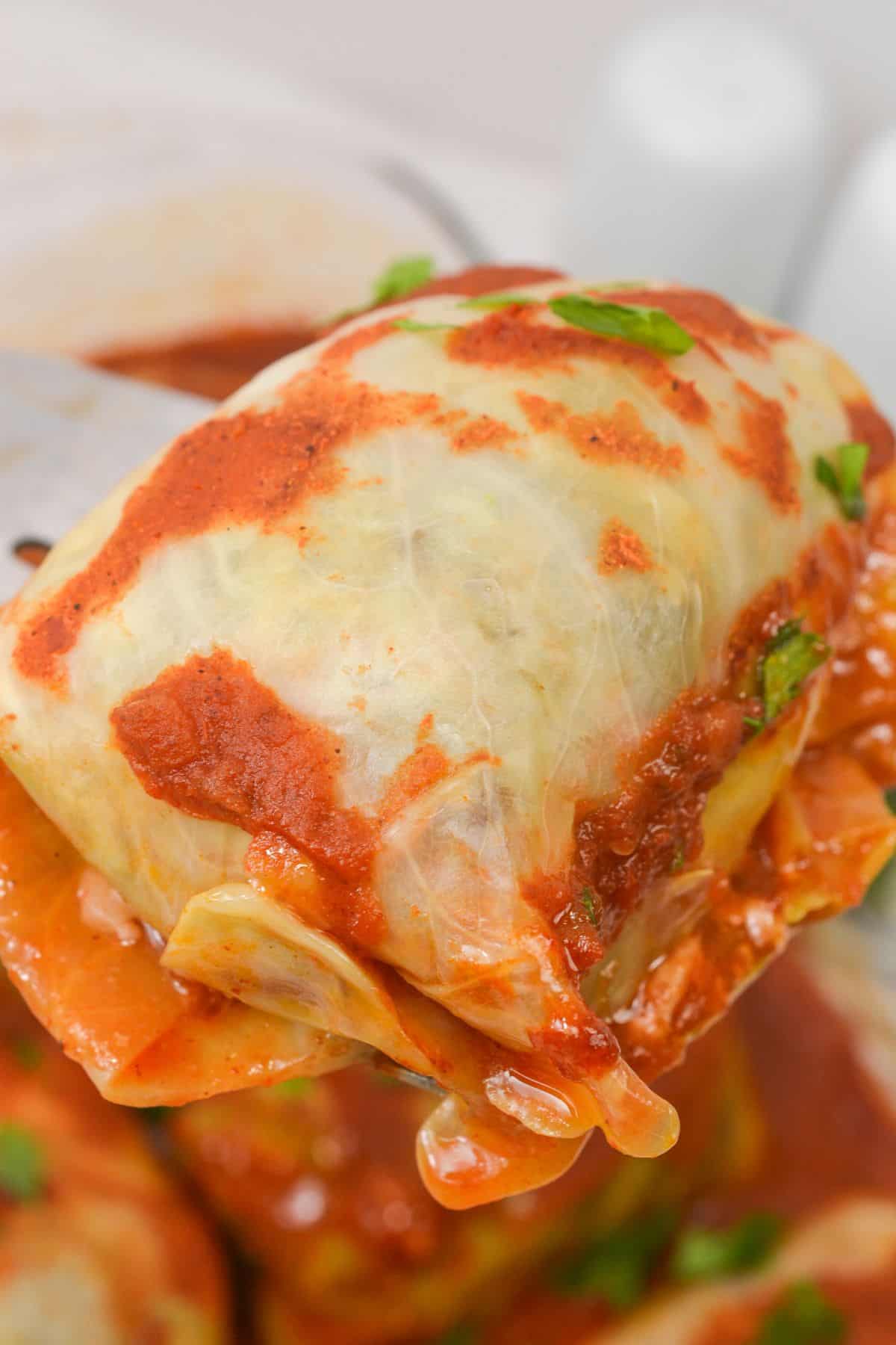 A serving utensil holds up an Easy Stuffed Cabbage Roll, cooked to perfection, covered in red tomato sauce and garnished with chopped herbs.