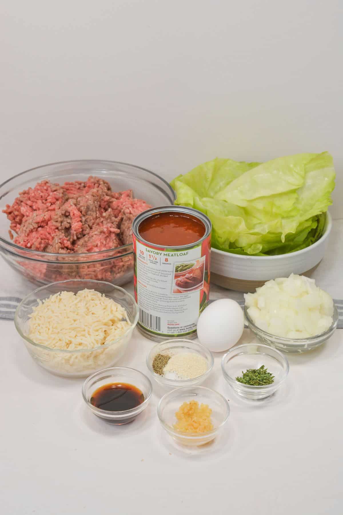 Assorted ingredients on a table, including ground beef, cabbage leaves, canned tomato sauce, rice, chopped onions, an egg, and seasonings in small bowls.