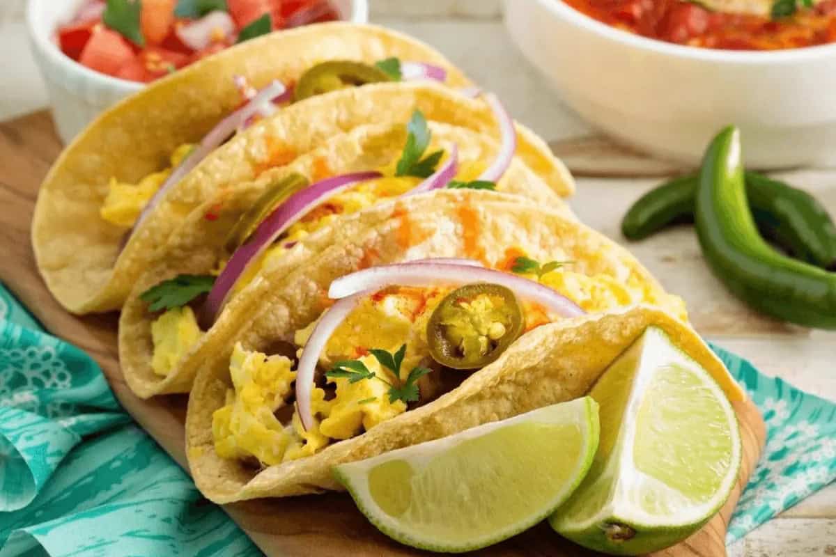 Three breakfast tacos filled with scrambled eggs, breakfast sausage, sliced red onion, jalapeños, and cilantro, garnished with lime wedges, are arranged on a wooden board.