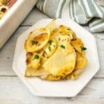 A serving of Potato Bake garnished with herbs on a white plate, next to a baking dish and a striped kitchen towel.