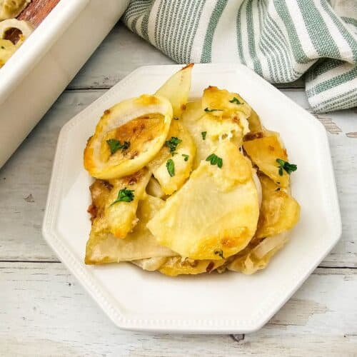 A serving of Potato Bake garnished with herbs on a white plate, next to a baking dish and a striped kitchen towel.