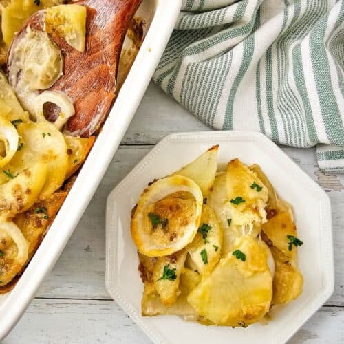 A serving of creamy Potato Bake with onions and herbs on a white plate, set beside a baking dish, wooden spoon, and a striped kitchen towel.