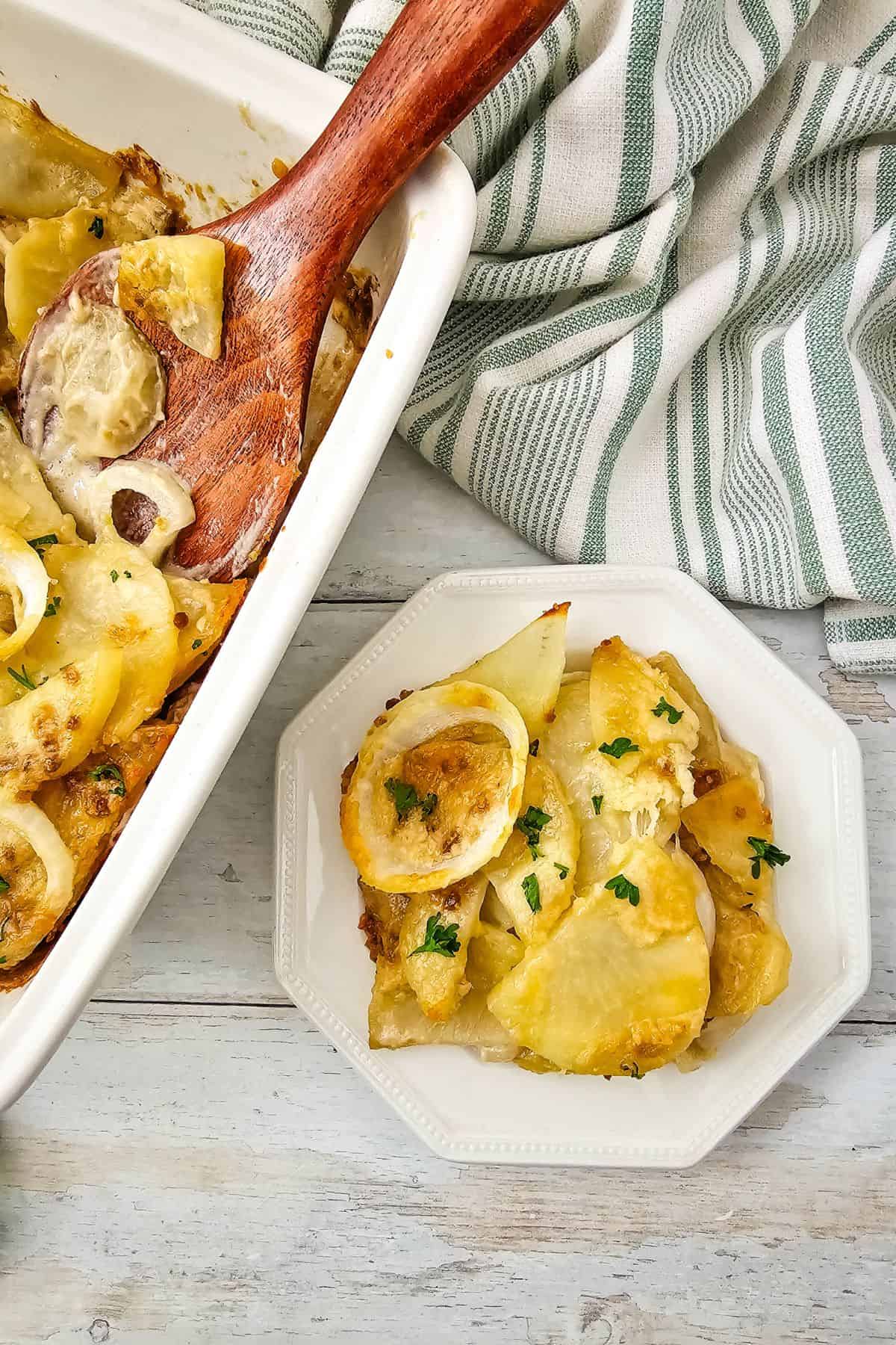 A serving of creamy Potato Bake with onions and herbs on a white plate, set beside a baking dish, wooden spoon, and a striped kitchen towel.