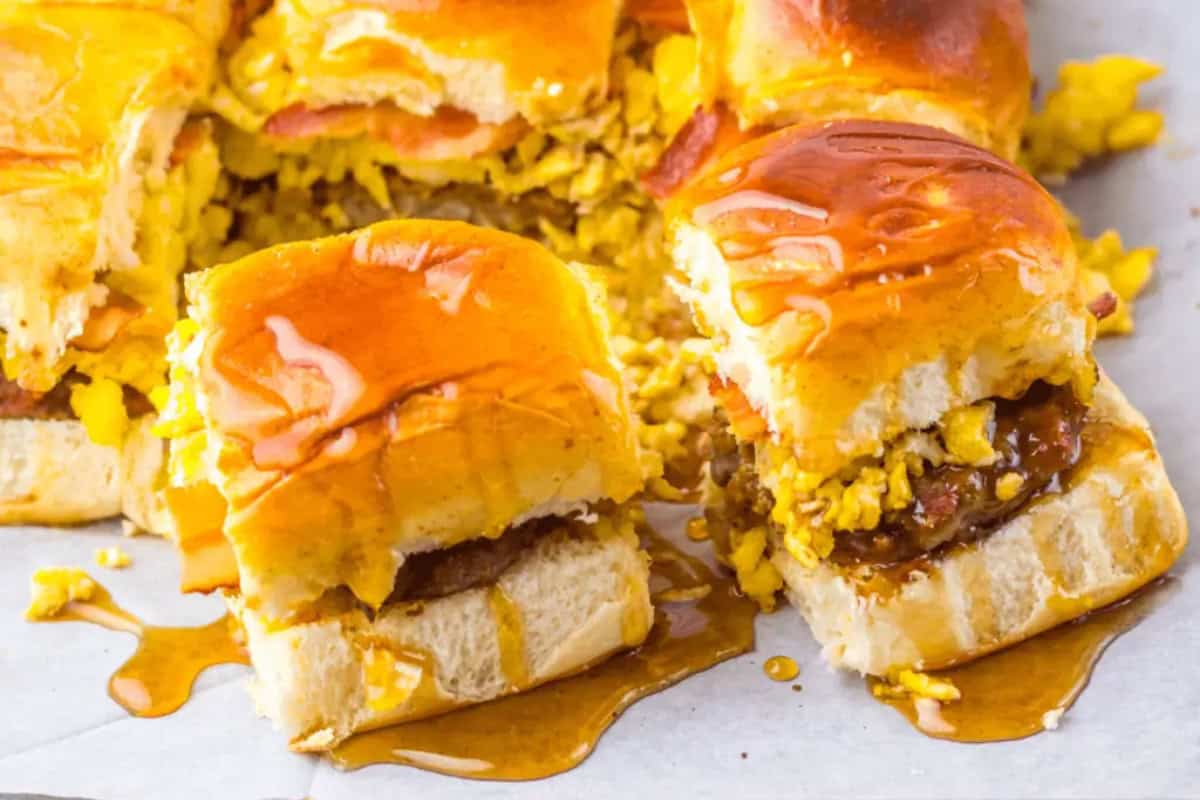 Breakfast sliders with scrambled eggs, breakfast sausage, and cheese on soft Sweet Hawaiian buns, drizzled with syrup, are arranged on parchment paper.