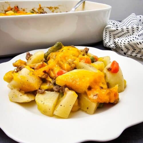 A serving of cheesy Hobo Casserole with potatoes, vegetables, and beef on a white plate; a casserole dish and patterned cloth are in the background.