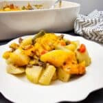 A white plate with a serving of Hobo Casserole featuring potatoes, vegetables, ground meat, and melted cheddar cheese, with a baking dish and spoon in the background.