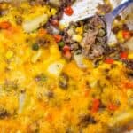A casserole dish containing a partially served cheesy pie with ground beef, mixed vegetables, and potatoes. A metal spoon rests in the dish.