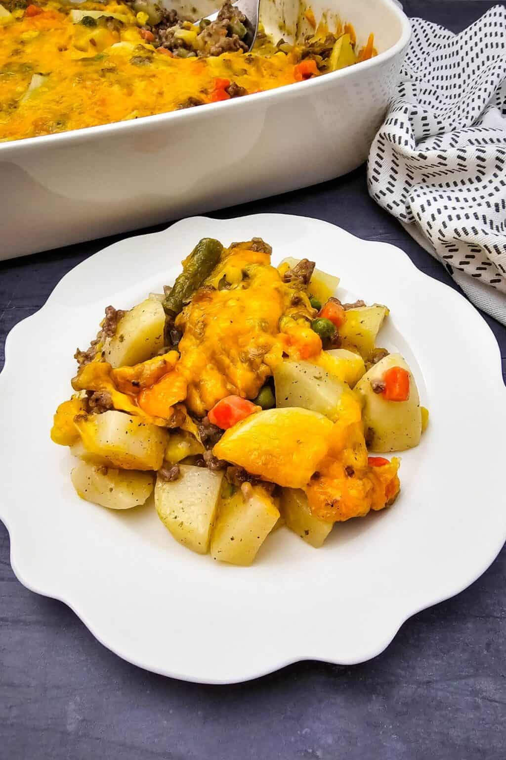 A serving of cheesy Hobo Casserole with beef, potatoes, and vegetables is plated on a white scalloped dish, with the casserole dish and a patterned napkin in the background.