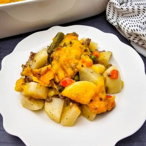 A serving of cheesy Hobo Casserole with beef, potatoes, and vegetables is plated on a white scalloped dish, with the casserole dish and a patterned napkin in the background.
