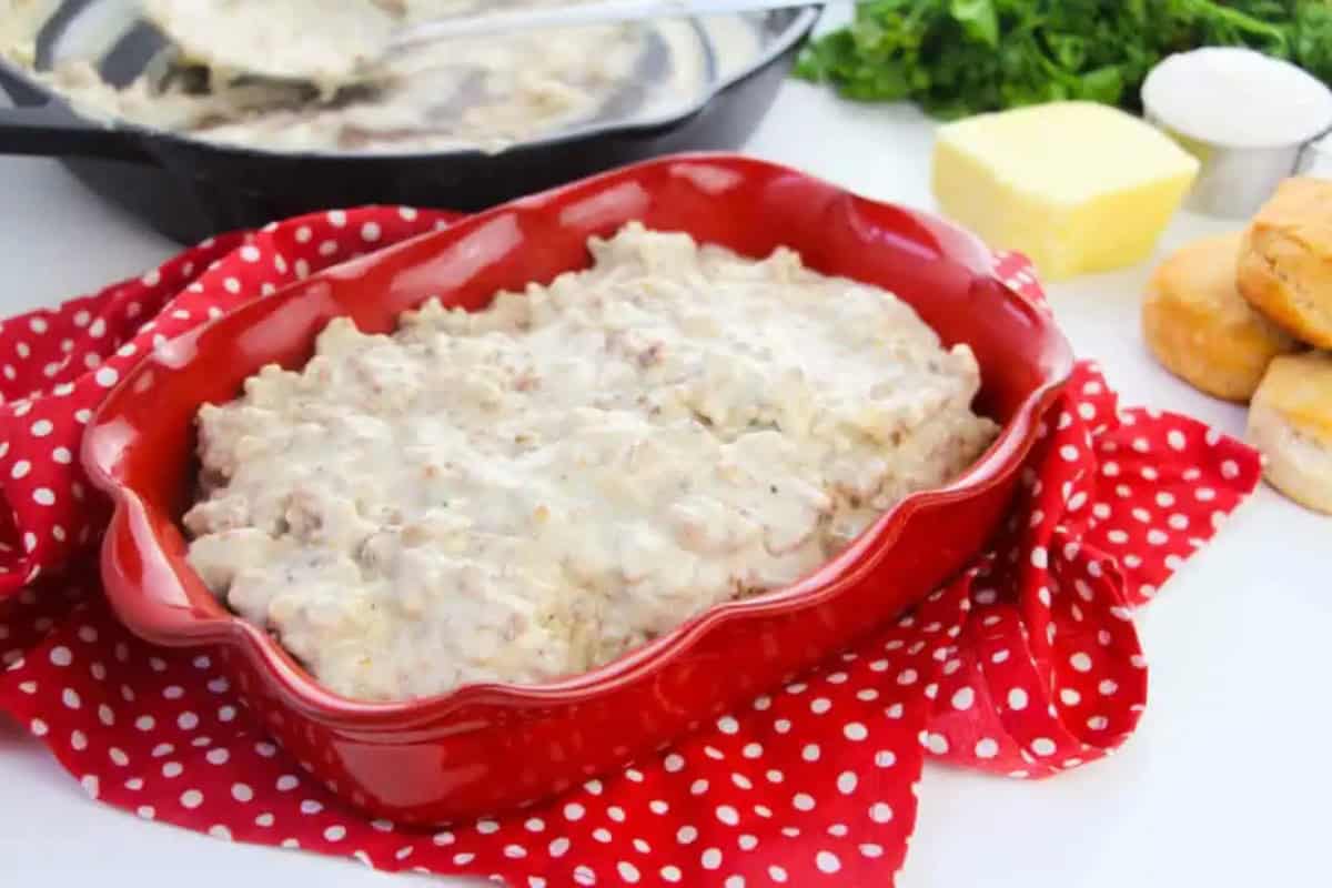 A red baking dish filled with sausage gravy sits on a red polka dot cloth, with biscuits, butter, and herbs nearby.