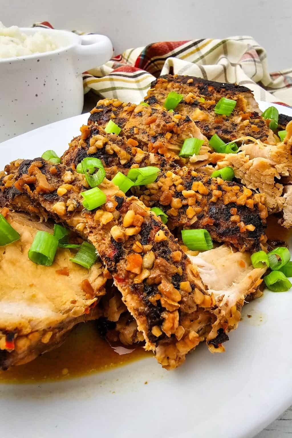 Crock Pot Pork Roast sliced and topped with chopped garlic and green onions on a white plate, with a bowl of rice in the background.