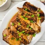 Crock Pot Pork Roast sliced and garnished with chopped green onions on a white oval plate, served alongside a bowl of mashed potatoes and a plaid cloth in the background.