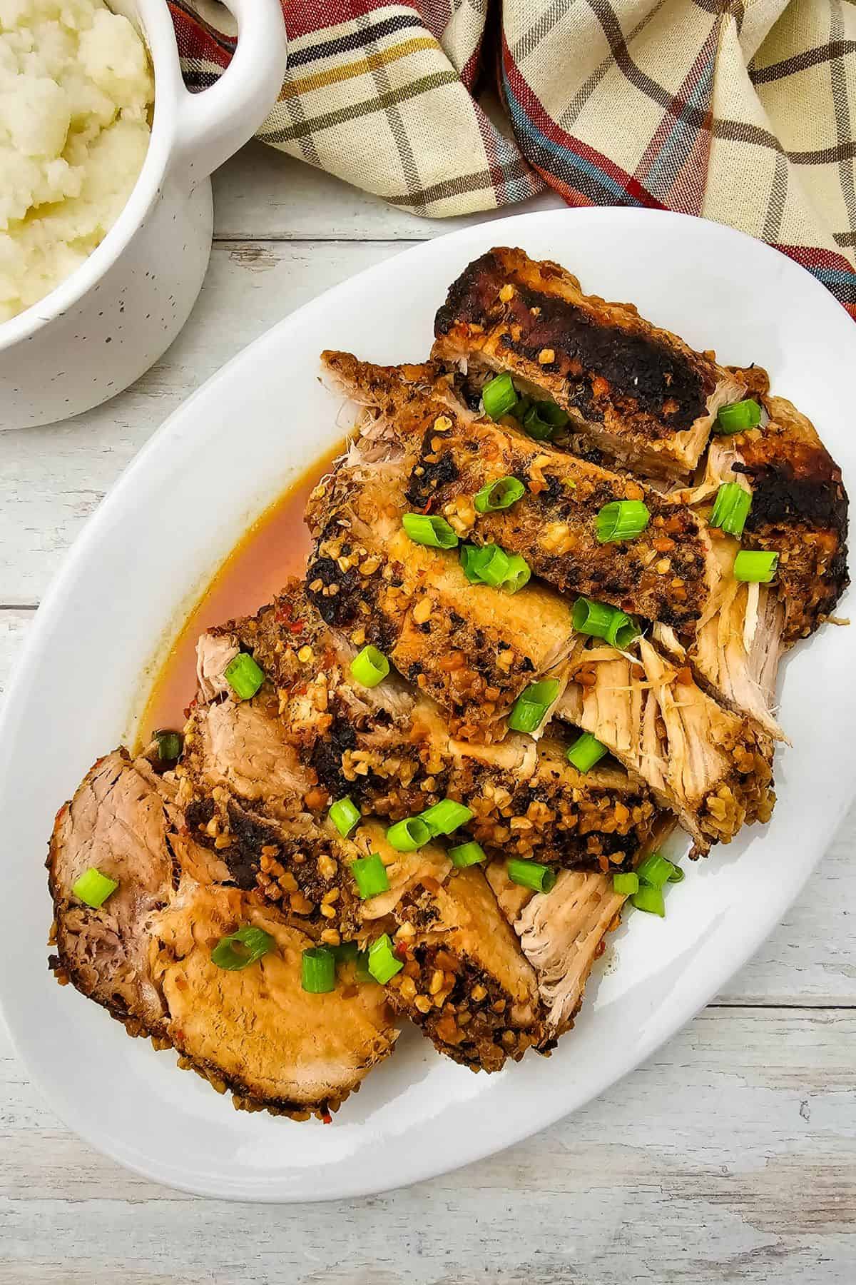Crock Pot Pork Roast sliced and garnished with chopped green onions on a white oval plate, served alongside a bowl of mashed potatoes and a plaid cloth in the background.