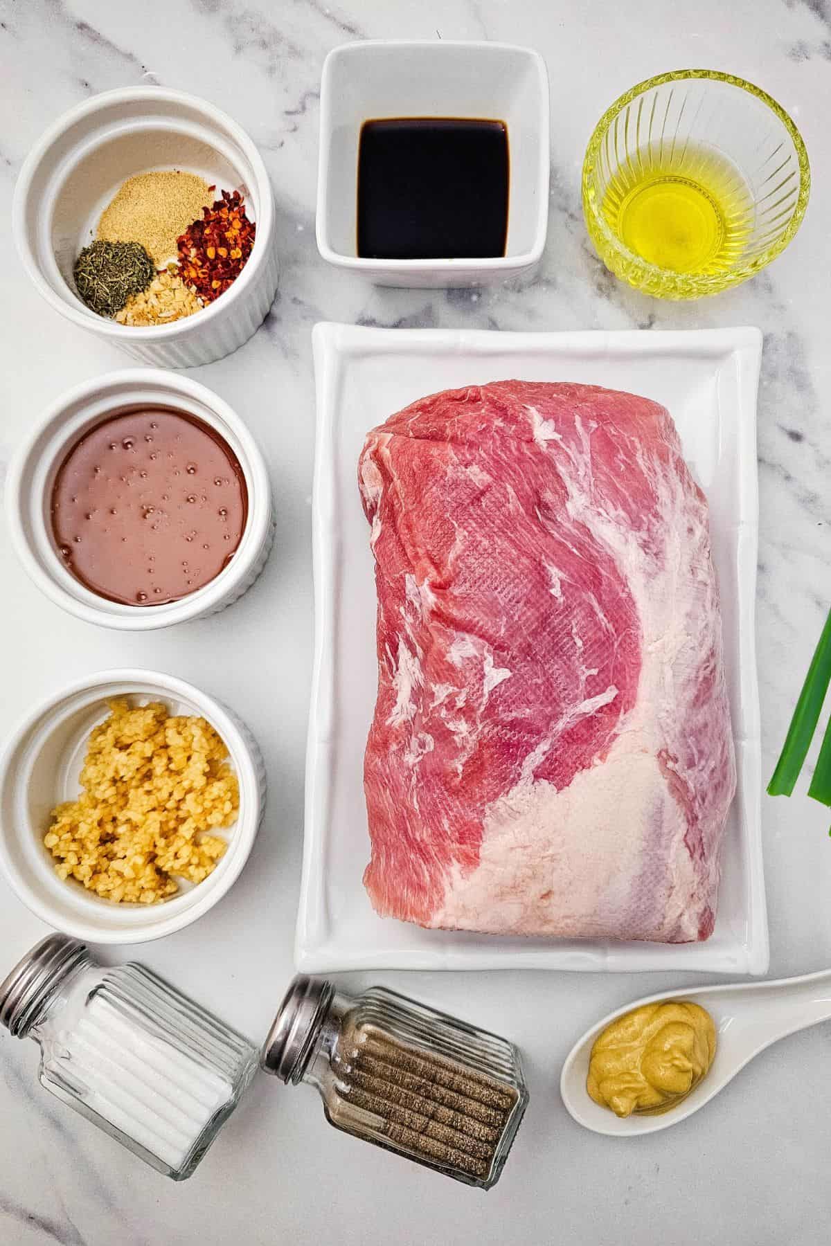 Raw pork shoulder on a white tray surrounded by bowls of minced garlic, spices, broth, mustard, oil, soy sauce, and salt and pepper—perfect ingredients for a flavorful Crock Pot Pork Roast on a marble surface.