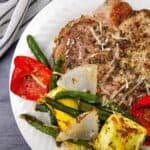 A white plate with Sheet Pan Pork Chops, grilled green beans, red bell pepper, yellow squash, and onions, garnished with shredded cheese, on a gray table with a striped cloth.