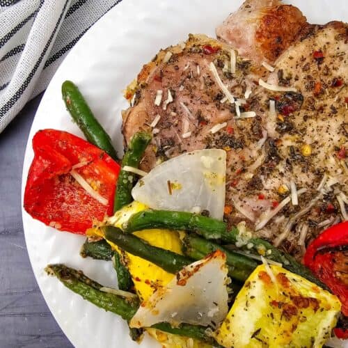 A white plate with Sheet Pan Pork Chops, grilled green beans, red bell pepper, yellow squash, and onions, garnished with shredded cheese, on a gray table with a striped cloth.