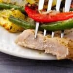 A fork holds a piece of cooked Sheet Pan Pork Chops and some roasted vegetables, including green beans and red peppers, above a white plate.