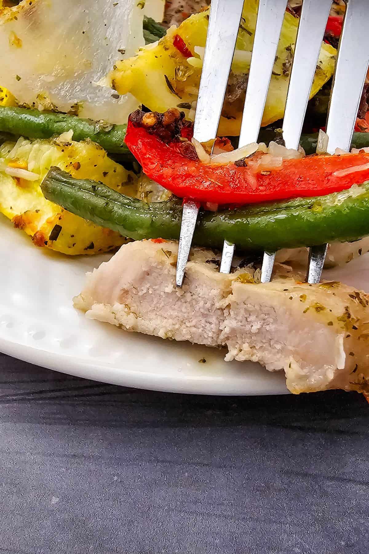 A fork holds a piece of cooked Sheet Pan Pork Chops and some roasted vegetables, including green beans and red peppers, above a white plate.