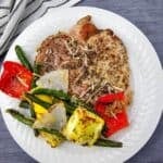 A seasoned Sheet Pan Pork Chop served on a white plate with grilled green beans, yellow squash, red bell pepper, and onion, presented on a dark surface beside a striped cloth.