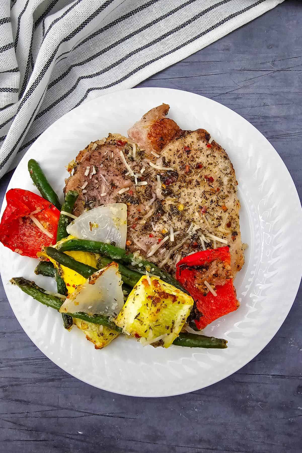 A seasoned Sheet Pan Pork Chop served on a white plate with grilled green beans, yellow squash, red bell pepper, and onion, presented on a dark surface beside a striped cloth.