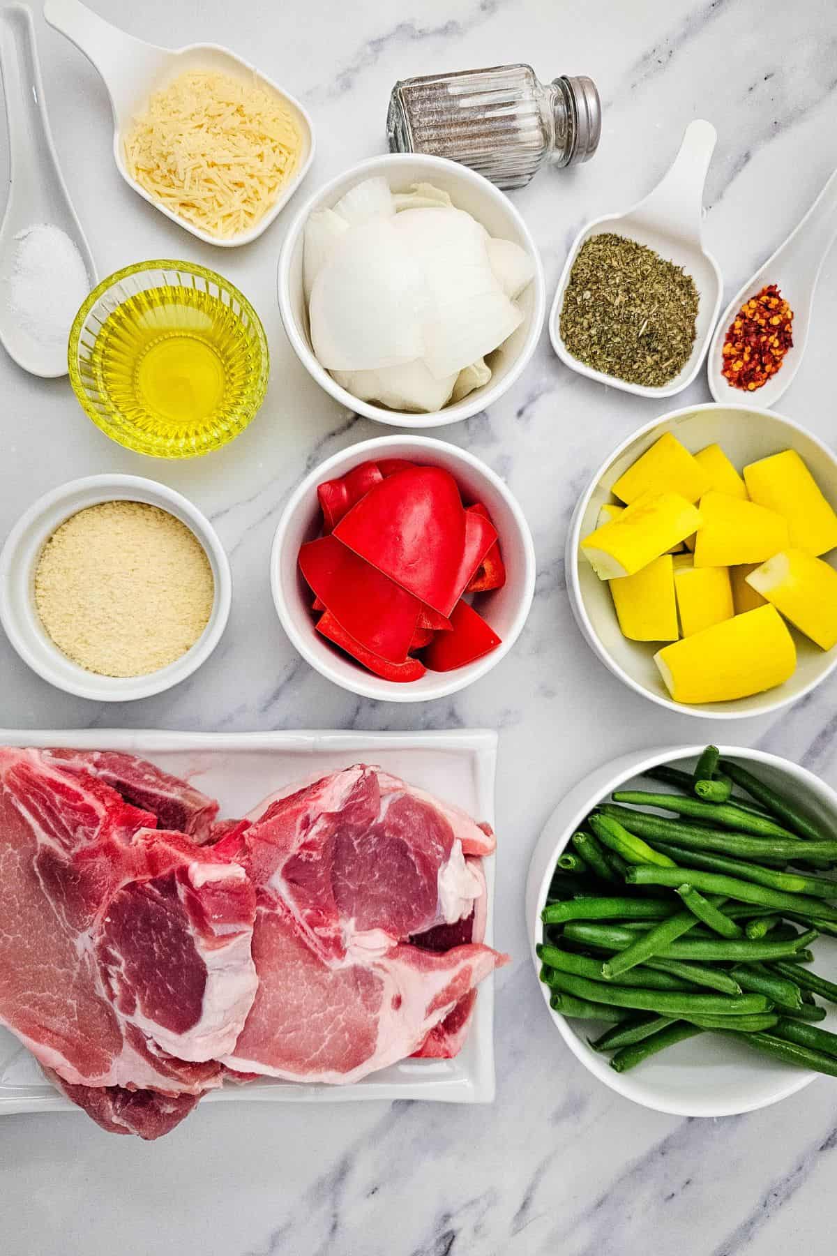 Meat, green beans, chopped red bell pepper, yellow squash, sliced onion, olive oil, grated cheese, spices, and seasonings are arranged in bowls on a white marble surface.