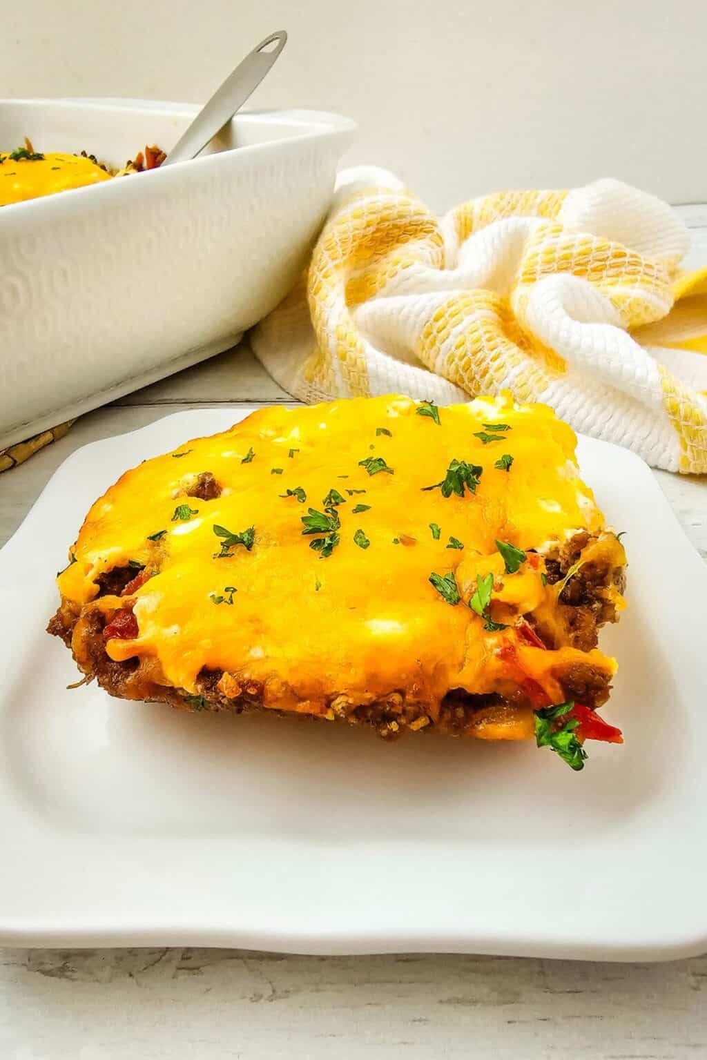 A slice of cheesy John Wayne Casserole with ground meat and vegetables sits on a white plate, with a baking dish and a yellow-and-white towel in the background.