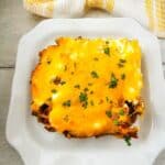 A serving of cheesy John Wayne Casserole topped with chopped parsley sits on a white square plate, with a yellow and white towel in the background.
