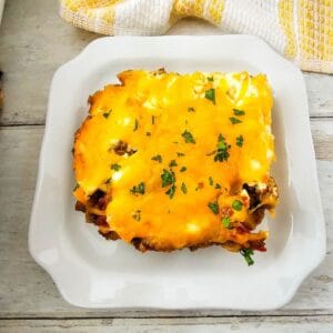 A square serving of cheesy John Wayne Casserole topped with melted cheddar and chopped herbs, on a white plate with a yellow and white towel in the background.