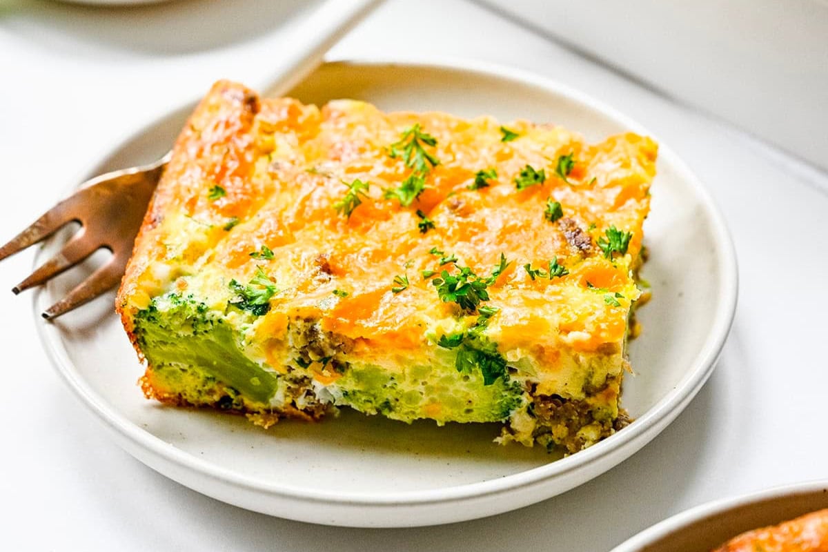 A slice of broccoli, egg and cheese casserole featuring breakfast sausage is garnished with chopped herbs and served on a white plate with a fork beside it.