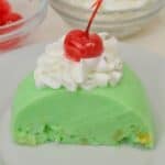 A slice of Lime Jello Salad, topped with whipped cream and a cherry, sits on a white plate, with bowls of cherries and whipped cream in the background.