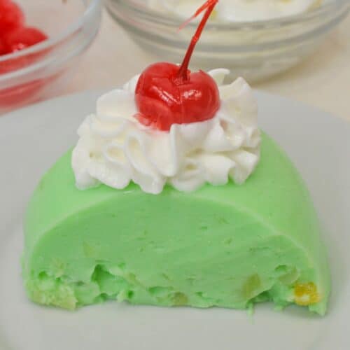 A slice of Lime Jello Salad, topped with whipped cream and a cherry, sits on a white plate, with bowls of cherries and whipped cream in the background.