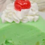 A slice of Lime Jello Salad topped with whipped cream and a maraschino cherry.
