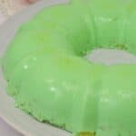 A green Lime Jello Salad mold ring sits on a white plate, with bowls of cherries and whipped cream in the background.