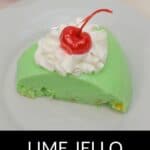 A slice of classic Lime Jello Salad topped with whipped cream and a maraschino cherry sits on a white plate.