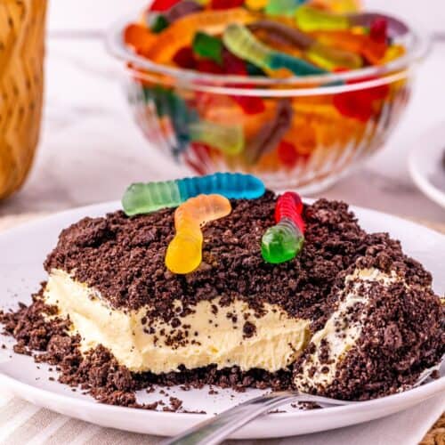 A dessert with a crumbly Oreo Dirt Cake topping and gummy worms on top, served on a white plate with a spoon; a bowl of gummy worms is in the background.