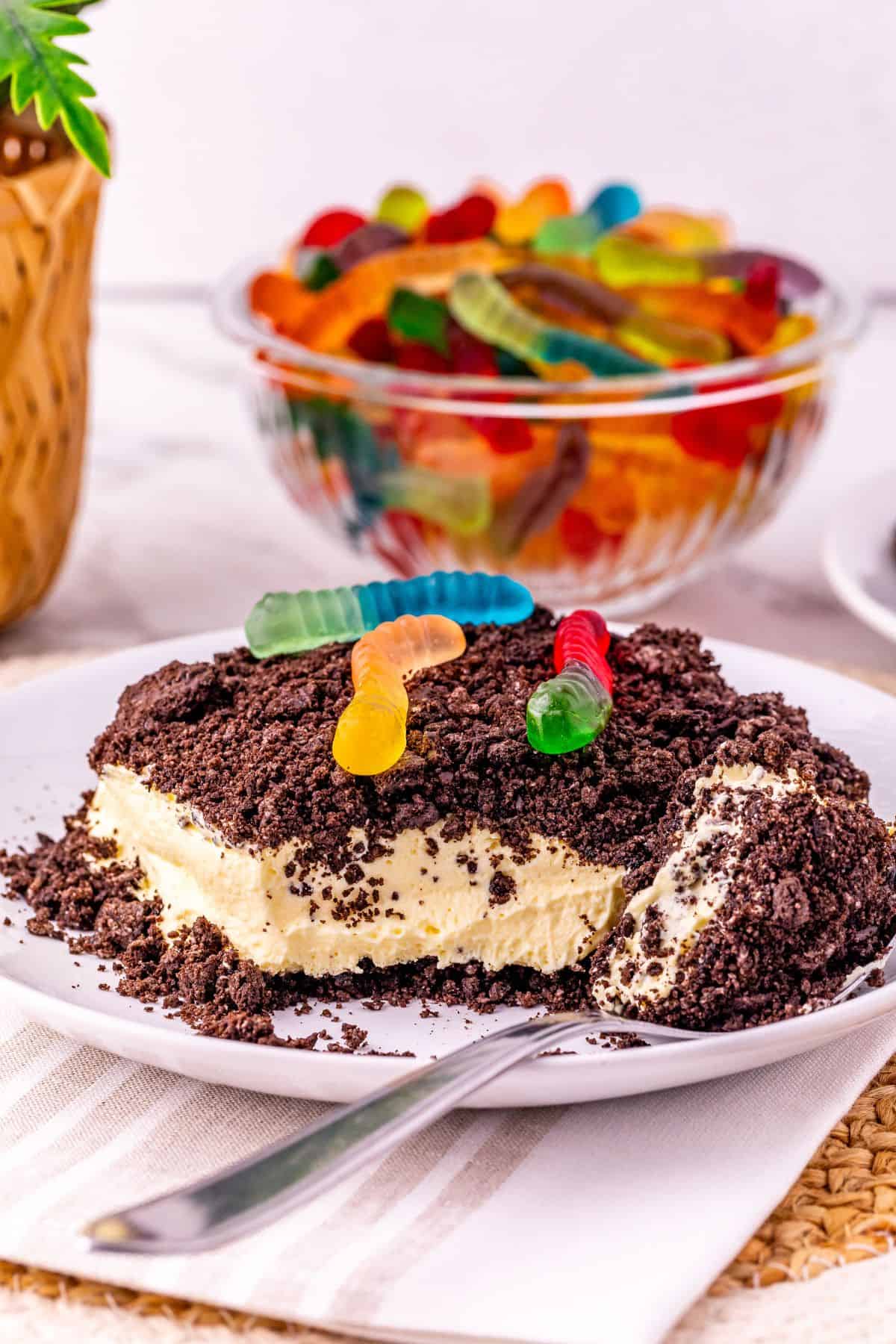 A dessert with a crumbly Oreo Dirt Cake topping and gummy worms on top, served on a white plate with a spoon; a bowl of gummy worms is in the background.