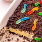 Rectangular dish with Oreo Dirt Cake, topped with crushed chocolate cookies and colorful gummy worms; a portion has been removed from one corner.