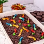 A rectangular dish filled with crumbled chocolate cookies and colorful gummy worms sits up front, with a bowl of gummy worms and a stack of sandwich cookies in the background.