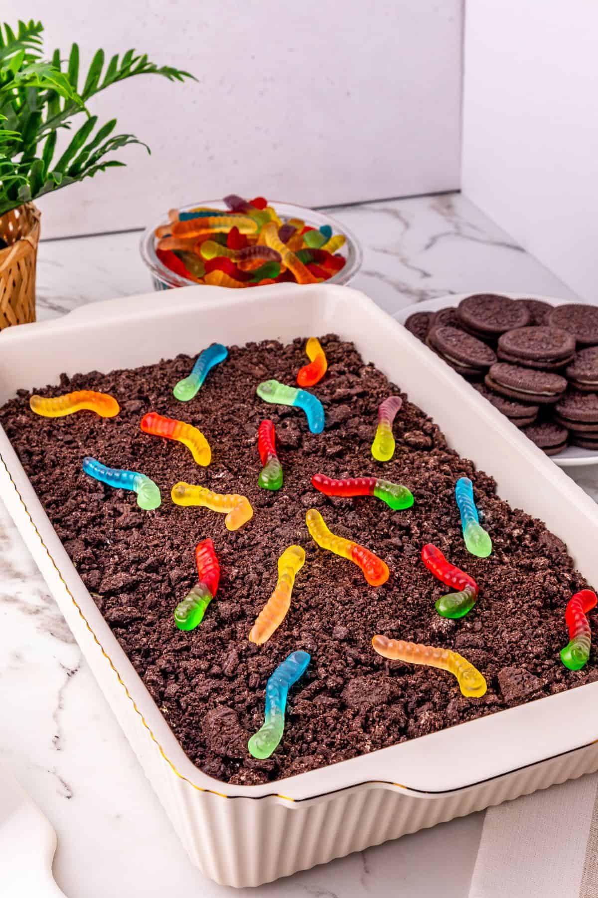 A rectangular dish filled with crumbled chocolate cookies and colorful gummy worms sits up front, with a bowl of gummy worms and a stack of sandwich cookies in the background.